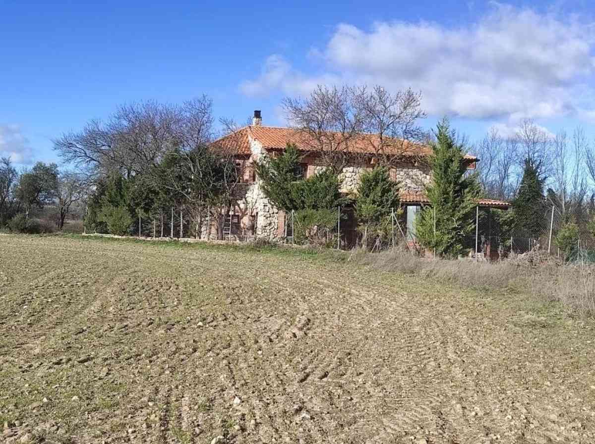 Casa rural con techo rojo junto a unos árboles, vista al otro lado de un campo arado bajo un cielo azul.