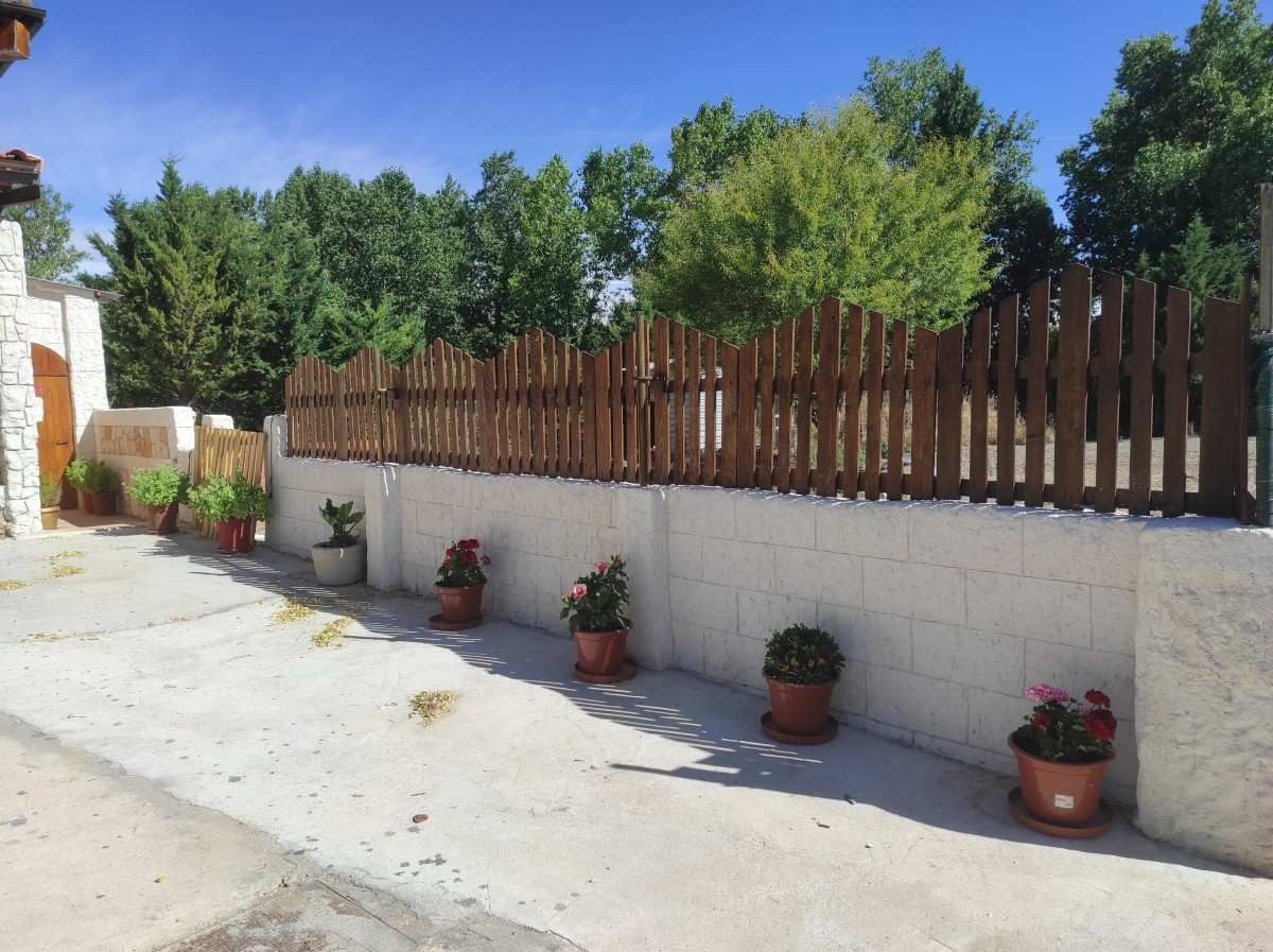 Muro blanco con macetas de flores a lo largo de un camino de entrada, rematado por una valla de madera bajo un cielo azul brillante.