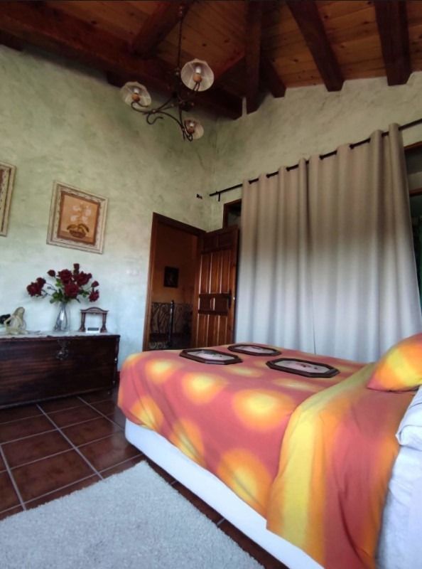 Acogedora habitación con techo de vigas de madera, colcha naranja, suelo de baldosas y gran ventana con cortinas.