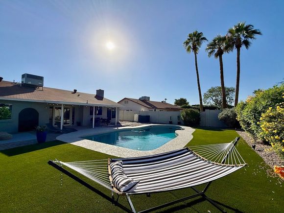 Backyard pool with striped lounge chair, green lawn, palm trees, and bright sun under a blue sky