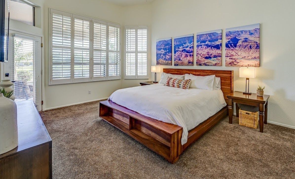 Bedroom with wooden bed, artwork, and large windows with shutters; tan carpet.