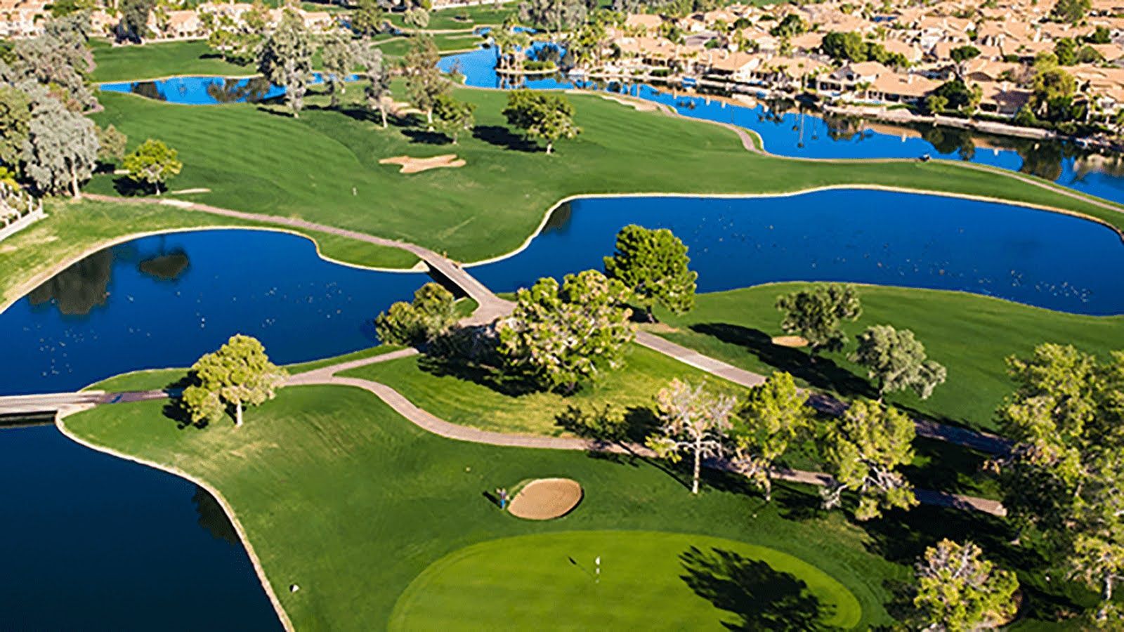 Aerial view of a green golf course with blue water features, trees, and houses in the background.