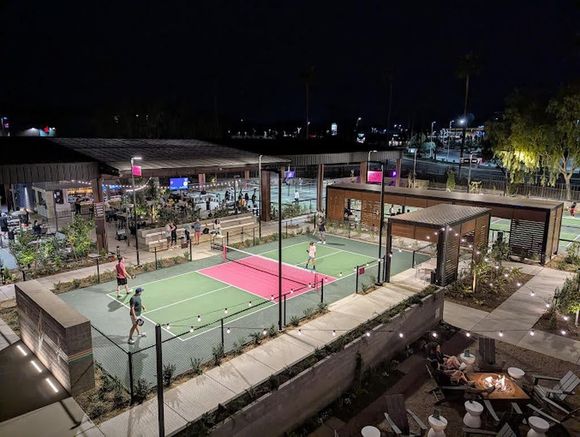Nighttime outdoor pickleball courts lit by floodlights, with players on green and pink courts and spectators nearby.