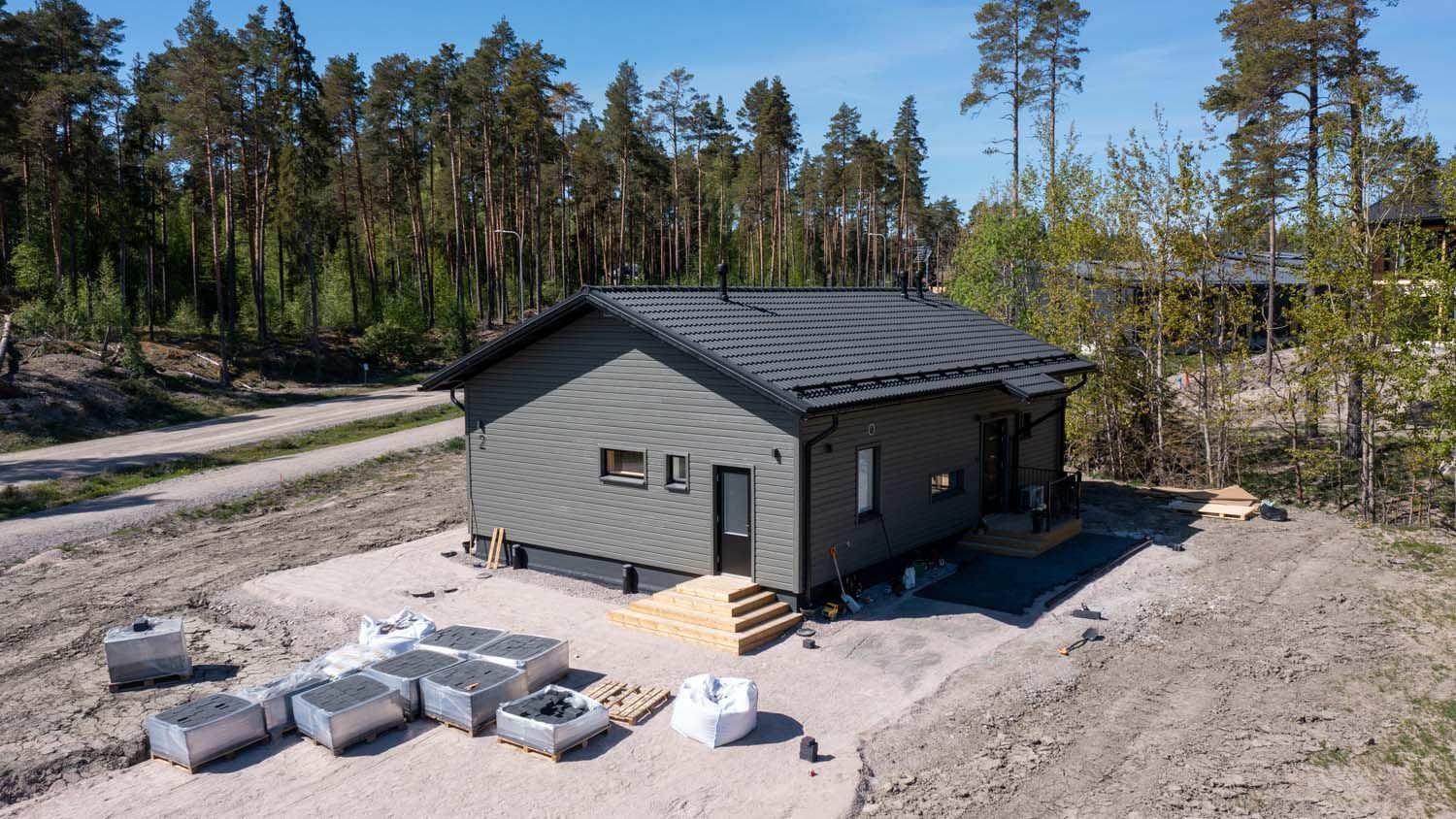 Harmaa talo rakenteilla soratontilla, puiden ja rakennusmateriaalien ympäröimänä.