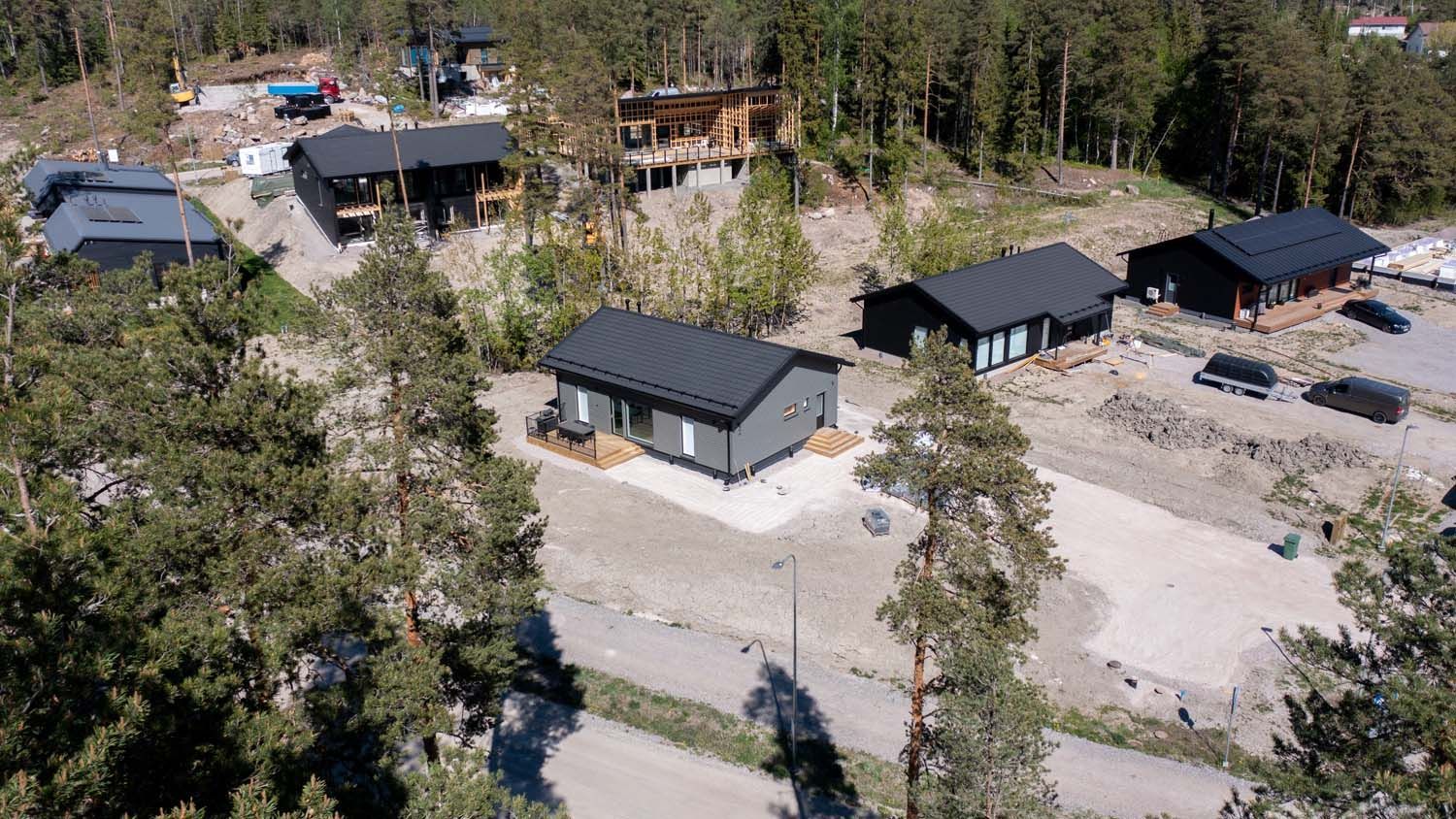 Ilmakuva useista tummakattoisista taloista hiekkatien varrella metsäisellä alueella.