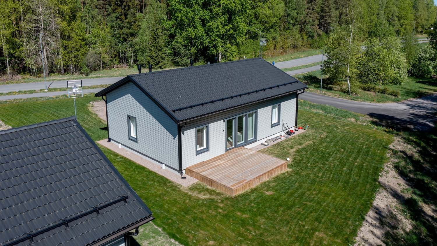 Pieni harmaa mökki, jossa on tumma katto ja patio nurmikolla tontilla, ylhäältä katsottuna tien ja puiden lähellä