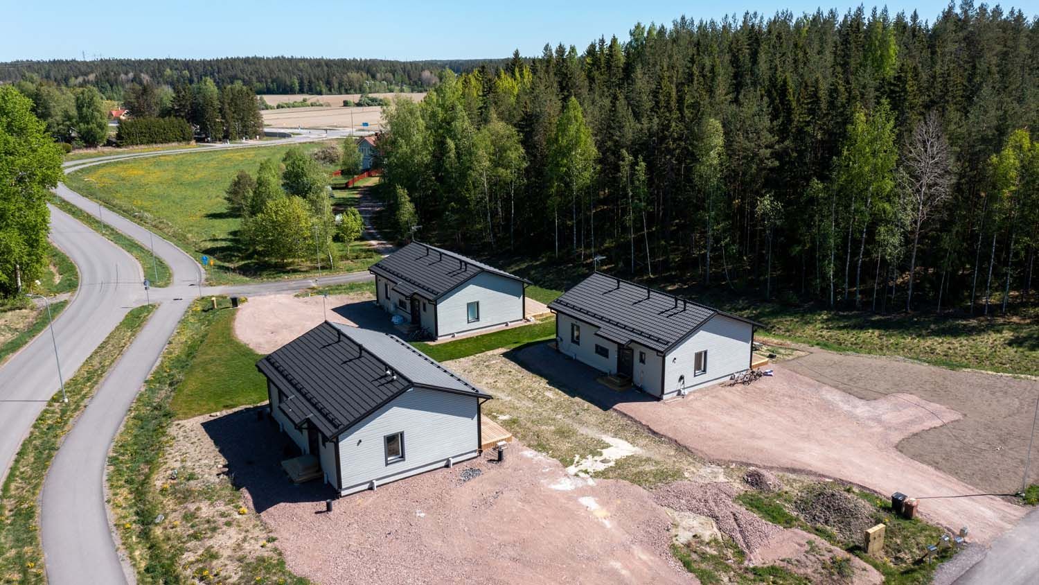 Ilmakuva kolmesta pienestä valkoisesta rakennuksesta maaseututien ja metsäaukeaman vieressä