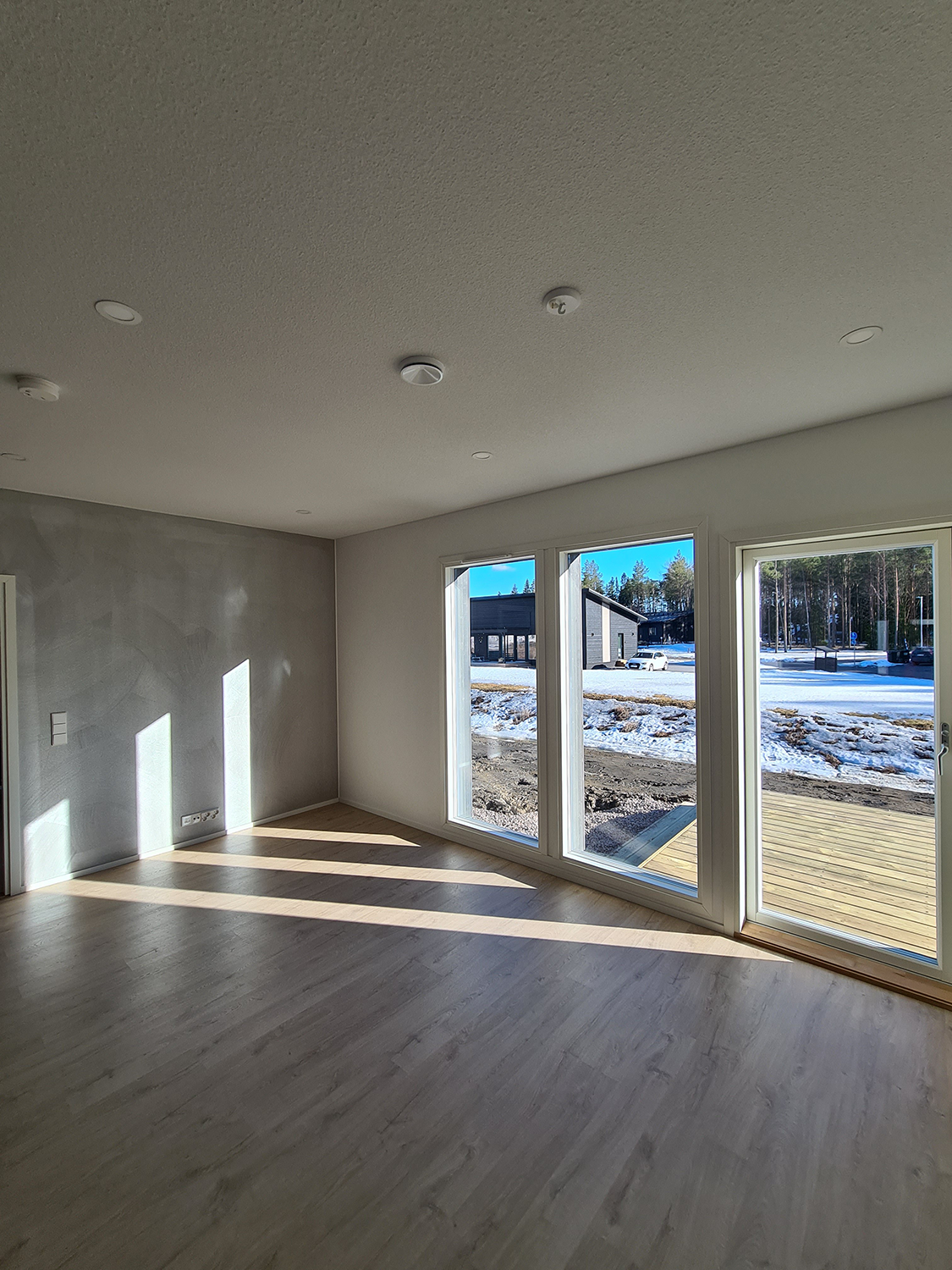 Tyhjä auringonvalossa oleva huone, jossa on suuret ikkunat, vaalea puulattia ja luminen ulkonäkymä
