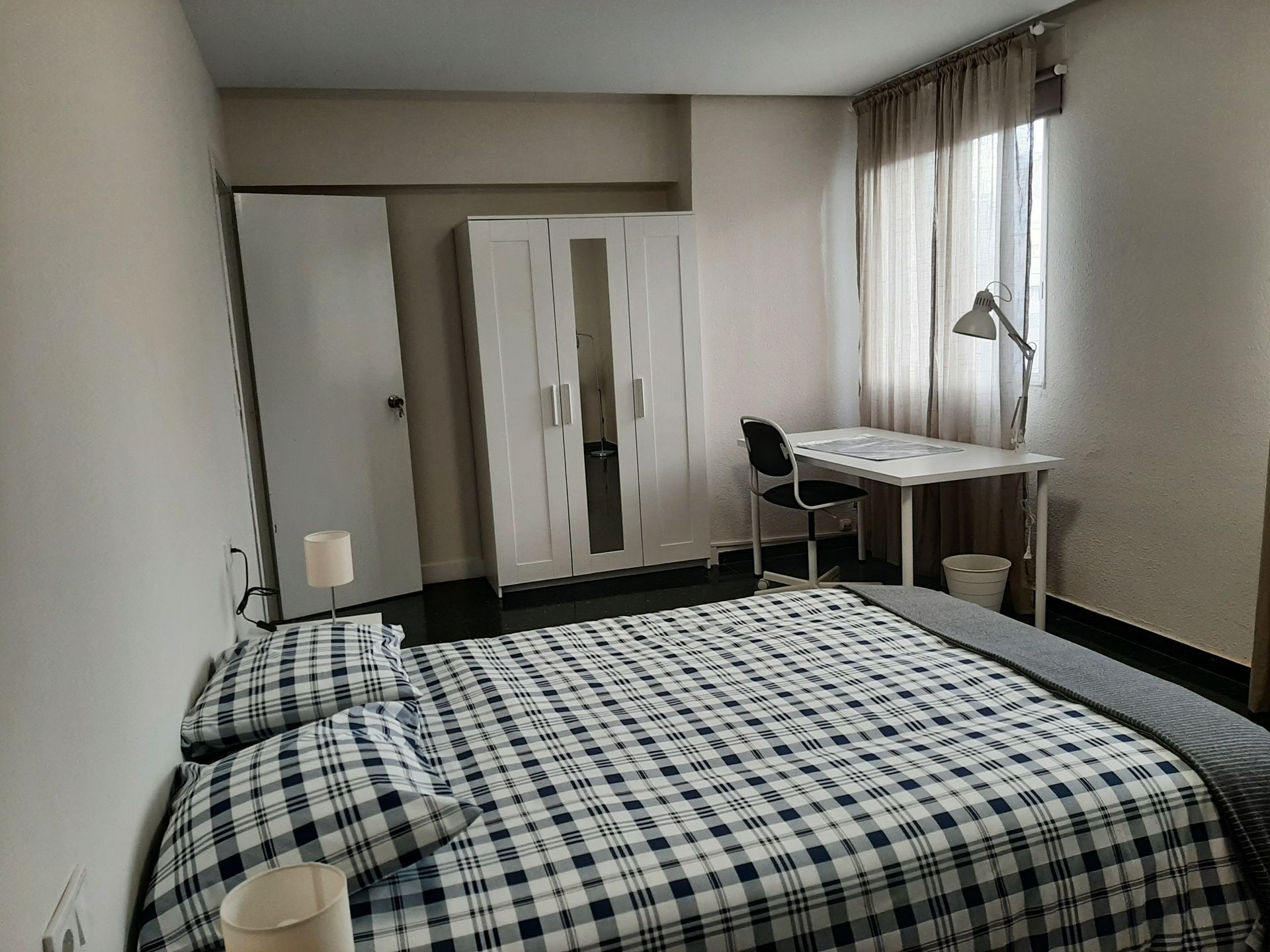 Dormitorio con cama, armario, escritorio y ventana con cortinas. Ropa de cama a cuadros azules.