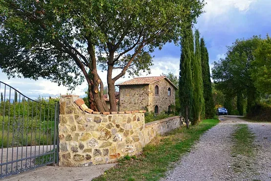Casa de piedra detrás de un muro de piedra y una puerta, con camino de grava y altos cipreses.