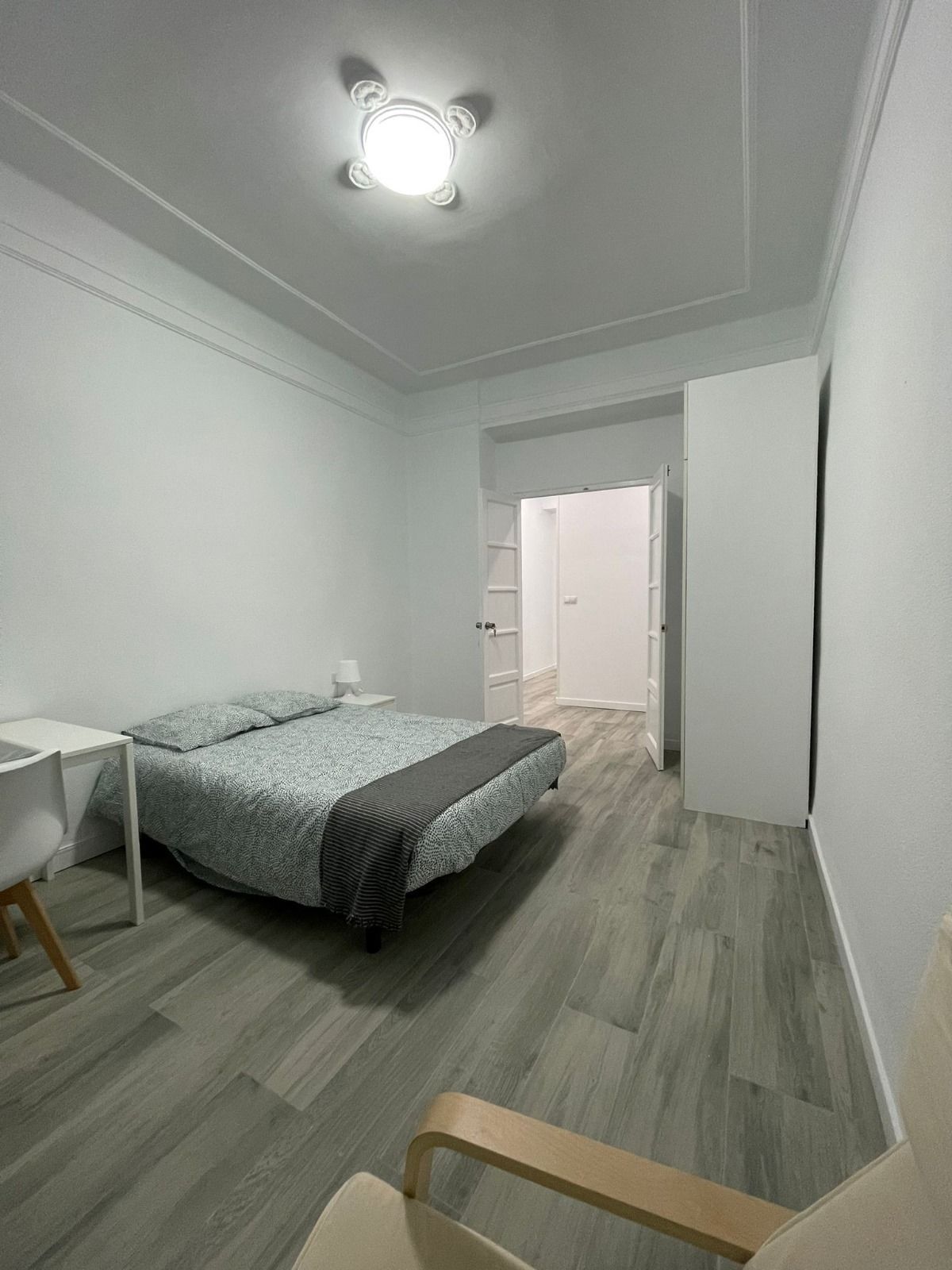 Dormitorio con cama, escritorio, silla y puerta abierta a un pasillo. Suelo gris imitación madera y paredes blancas.