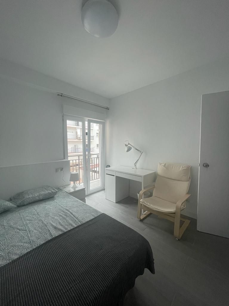 Dormitorio con cama, escritorio, silla y balcón. Paredes blancas, suelo gris.