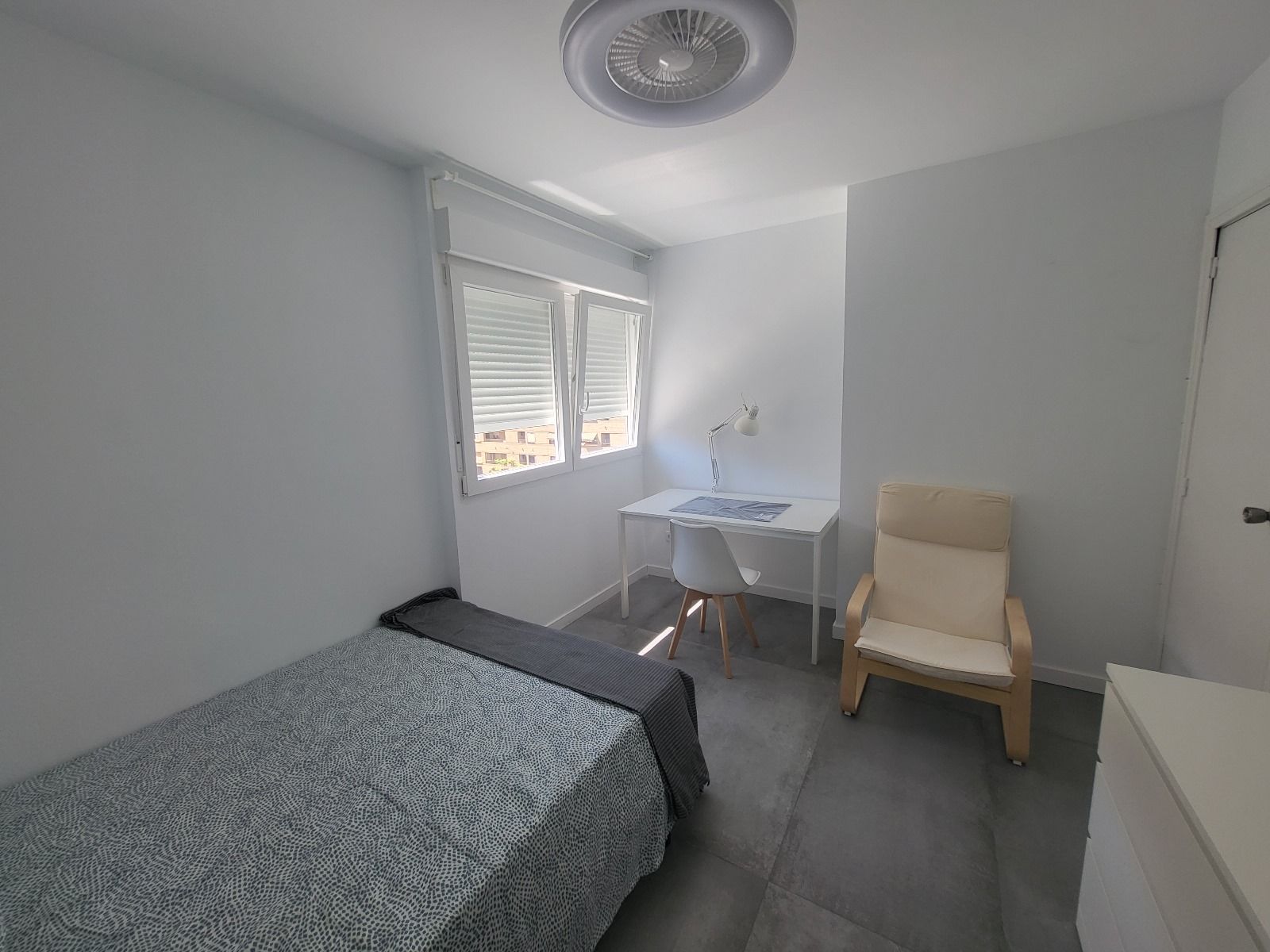 Dormitorio pequeño y blanco con una cama individual, escritorio, silla y ventana.