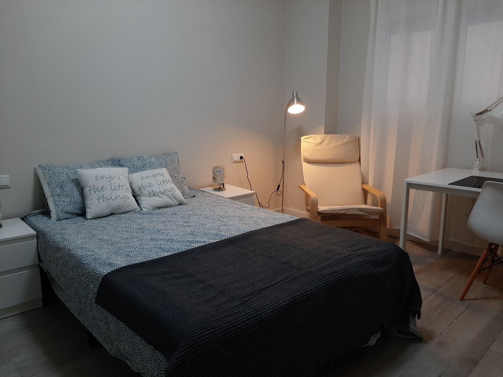 Dormitorio con cama, silla, escritorio y lámpara. Paredes blancas, ropa de cama azul y gris, luz natural.