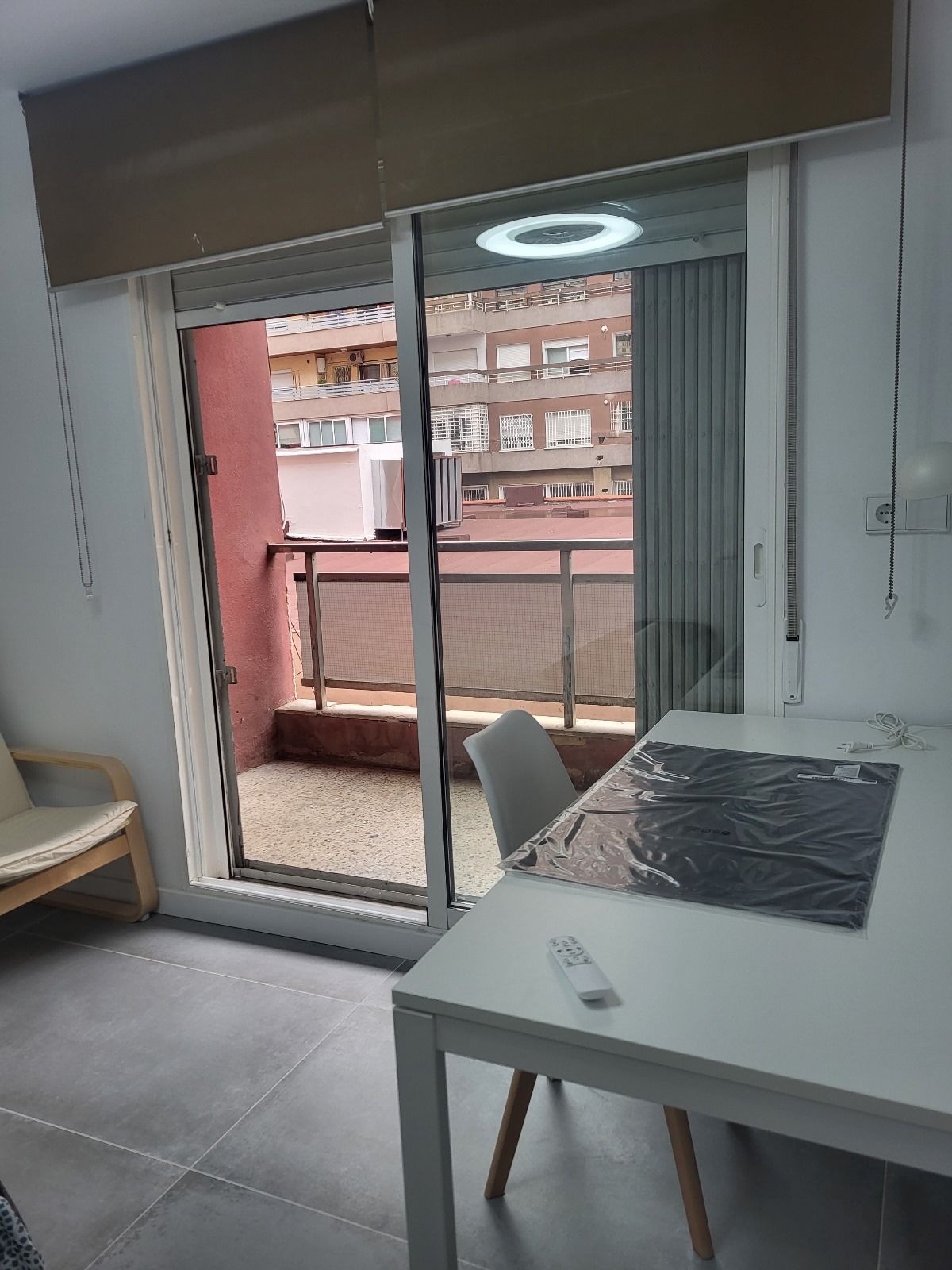 Interior con acceso al balcón: mesa, silla, puerta corrediza abierta, vista a la ciudad, persianas marrones y luz.