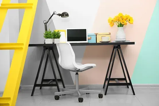 Escritorio con computadora portátil, silla, lámpara y flores en una habitación con pintura de pared colorida y una escalera amarilla.