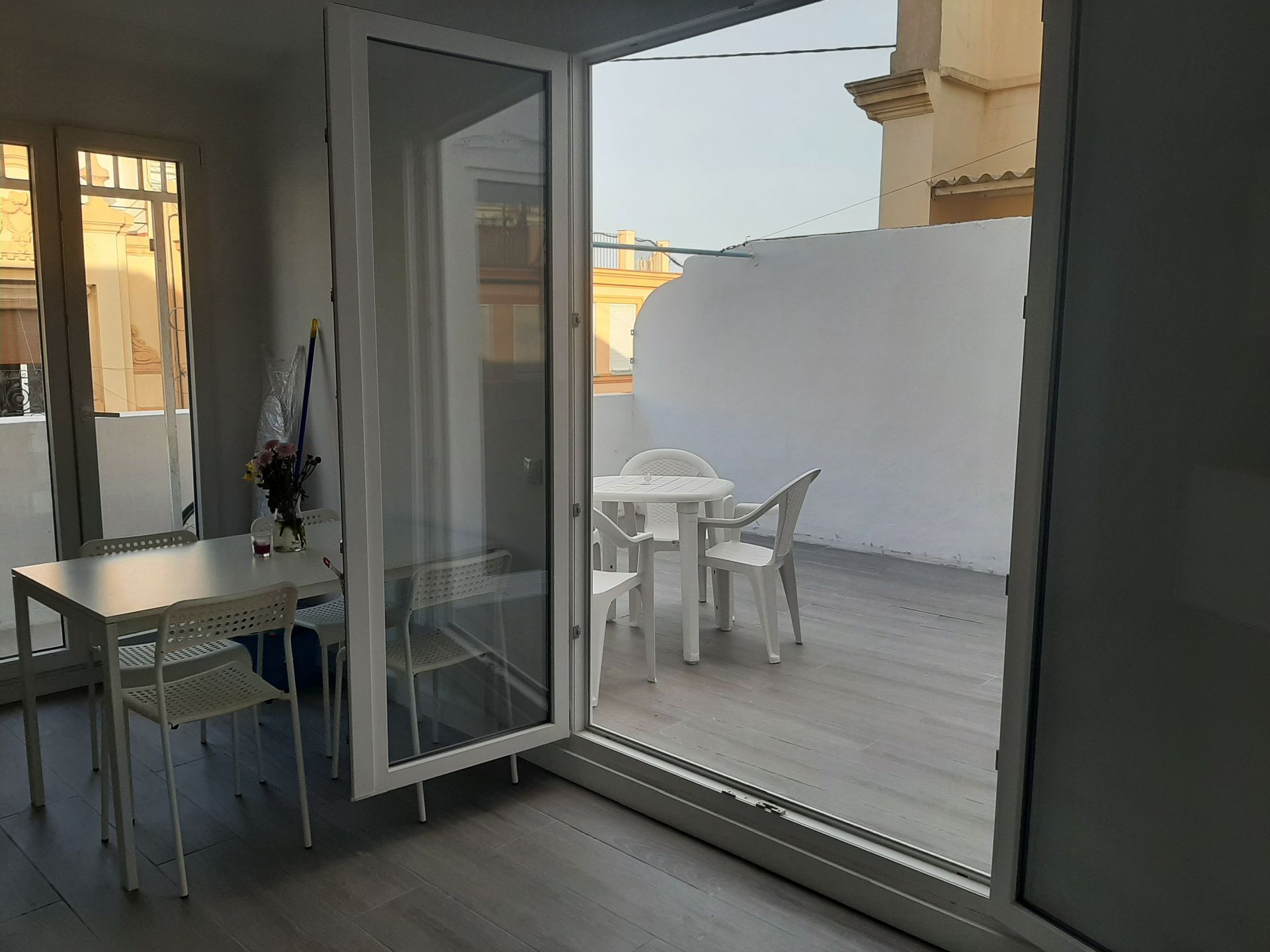 Puerta abierta a un pequeño patio con mesa y sillas blancas; una mesa más pequeña adentro con sillas.