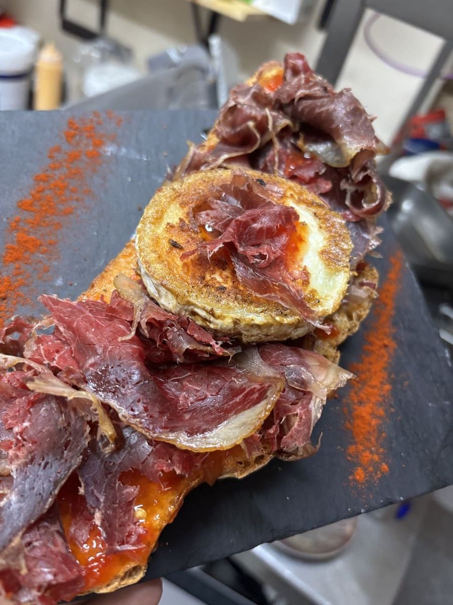 Plato de carne sellada, cubierto con tocino crujiente y condimento rojo, servido en una tabla de pizarra negra.