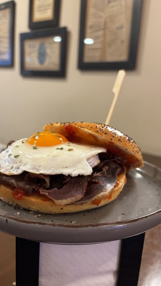 Sándwich de desayuno con un huevo frito encima, servido en un plato gris, con un cuadro borroso al fondo.