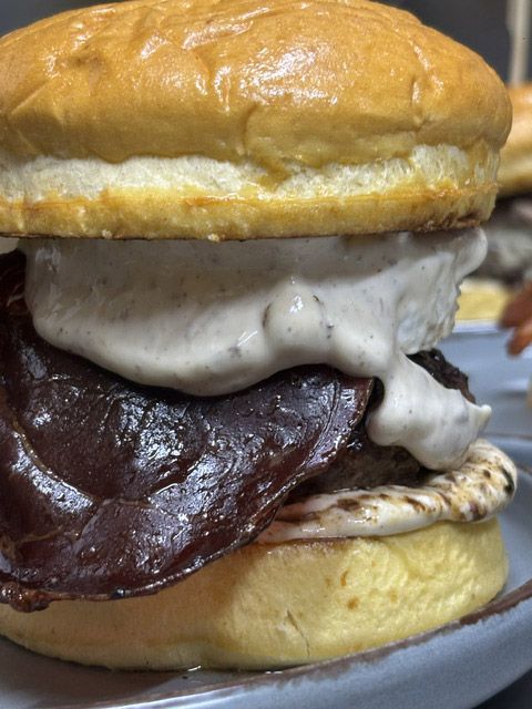 Hamburguesa alta con pan brillante, salsa blanca espesa y una hamburguesa de carne oscura servida en un plato.
