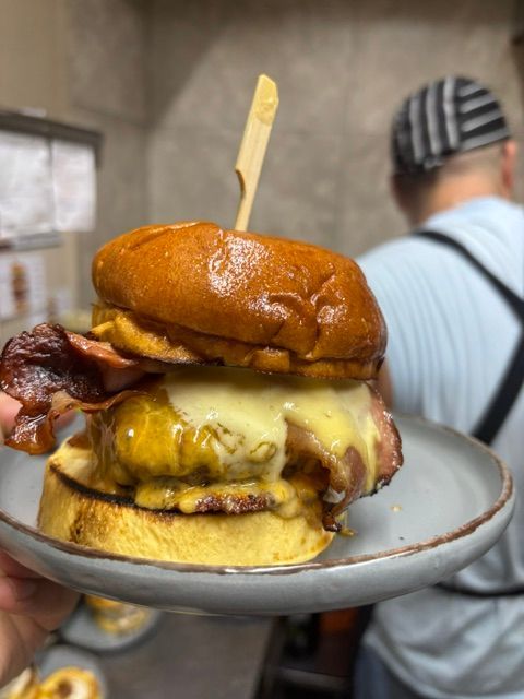 Hamburguesa con queso fundido y beicon en un plato, sostenida frente a una persona en una cocina.