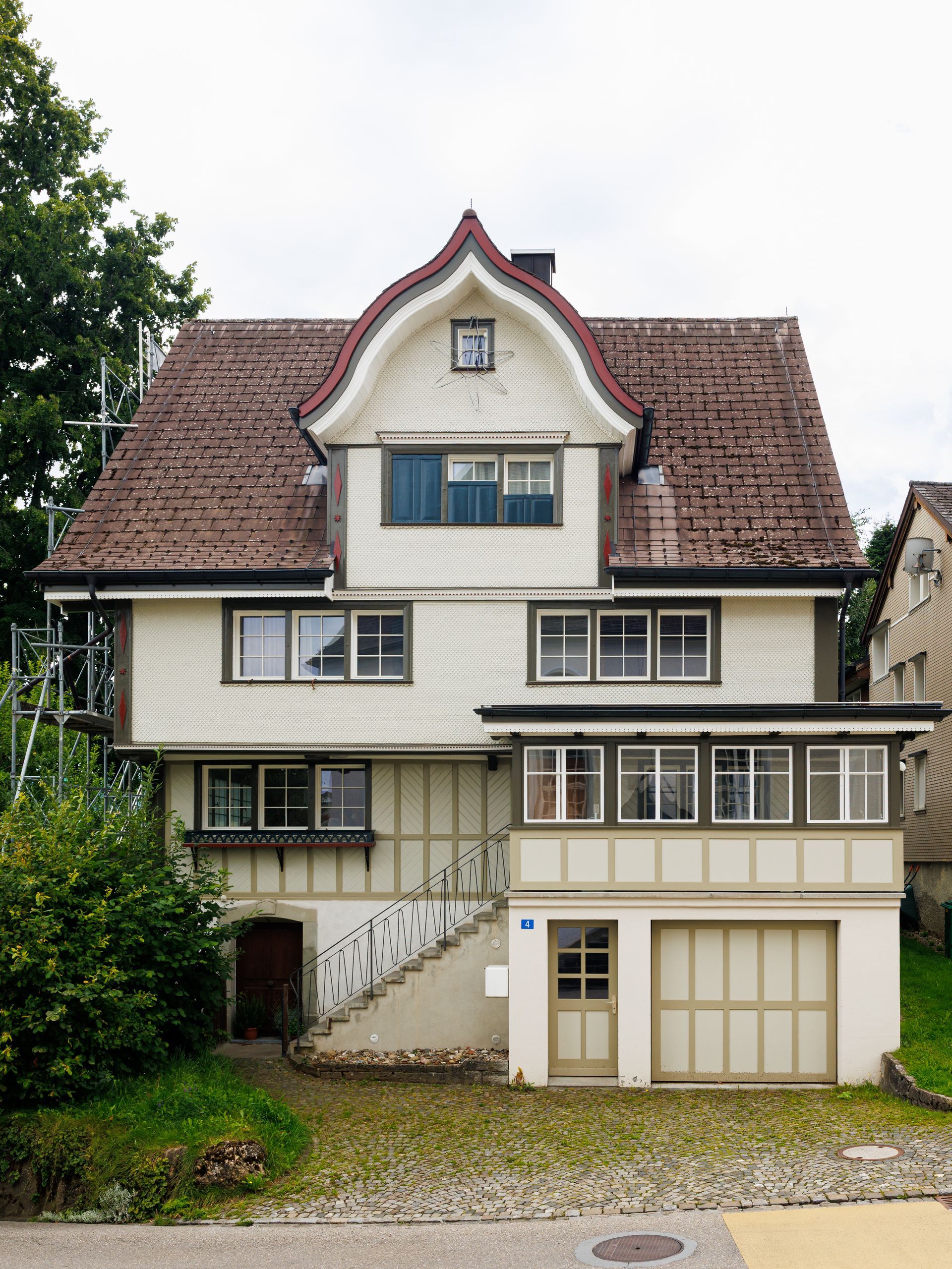 Zweistöckiges Haus mit rotem Ziegeldach und weißer Fassade; eine Garage und eine Veranda sind sichtbar.
