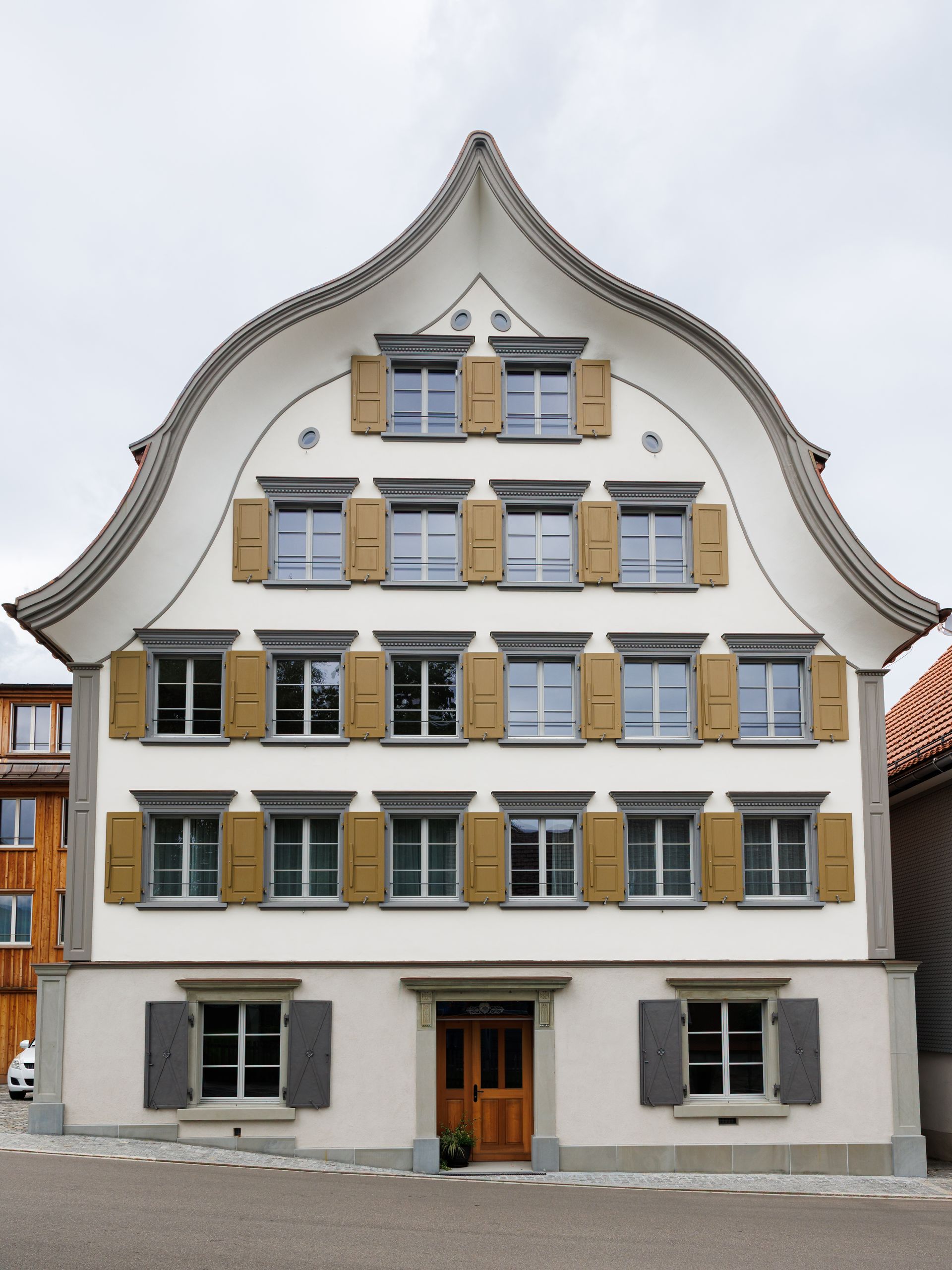 Cremefarbenes mehrstöckiges Gebäude mit gewölbtem Dach, hellbraunen Fensterläden und Holztür in einer Kopfsteinpflasterstraße.