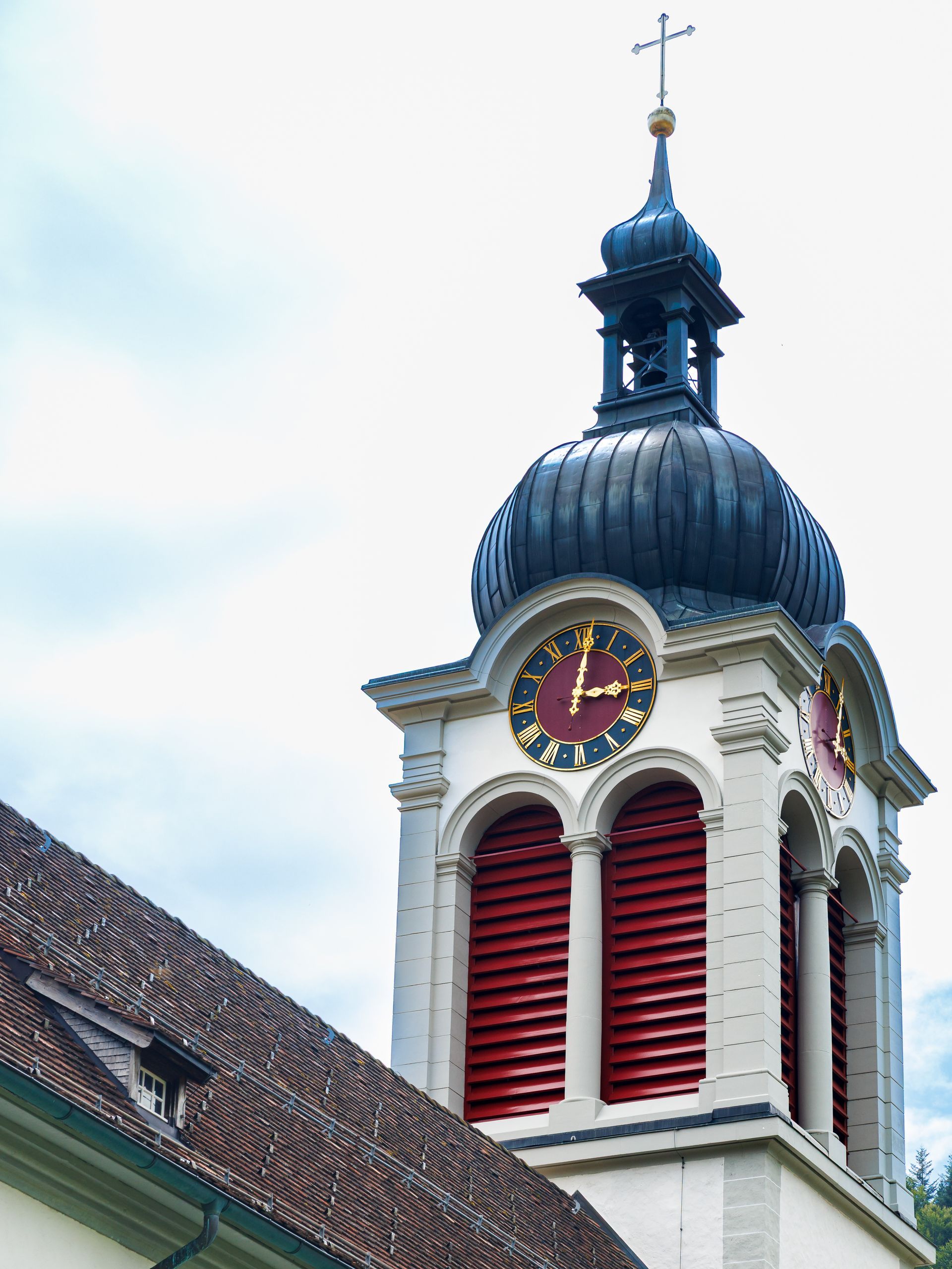Uhrturm mit dunkelblauer Kuppel, Zifferblatt, roten Fensterläden und Kreuz auf der Spitze.