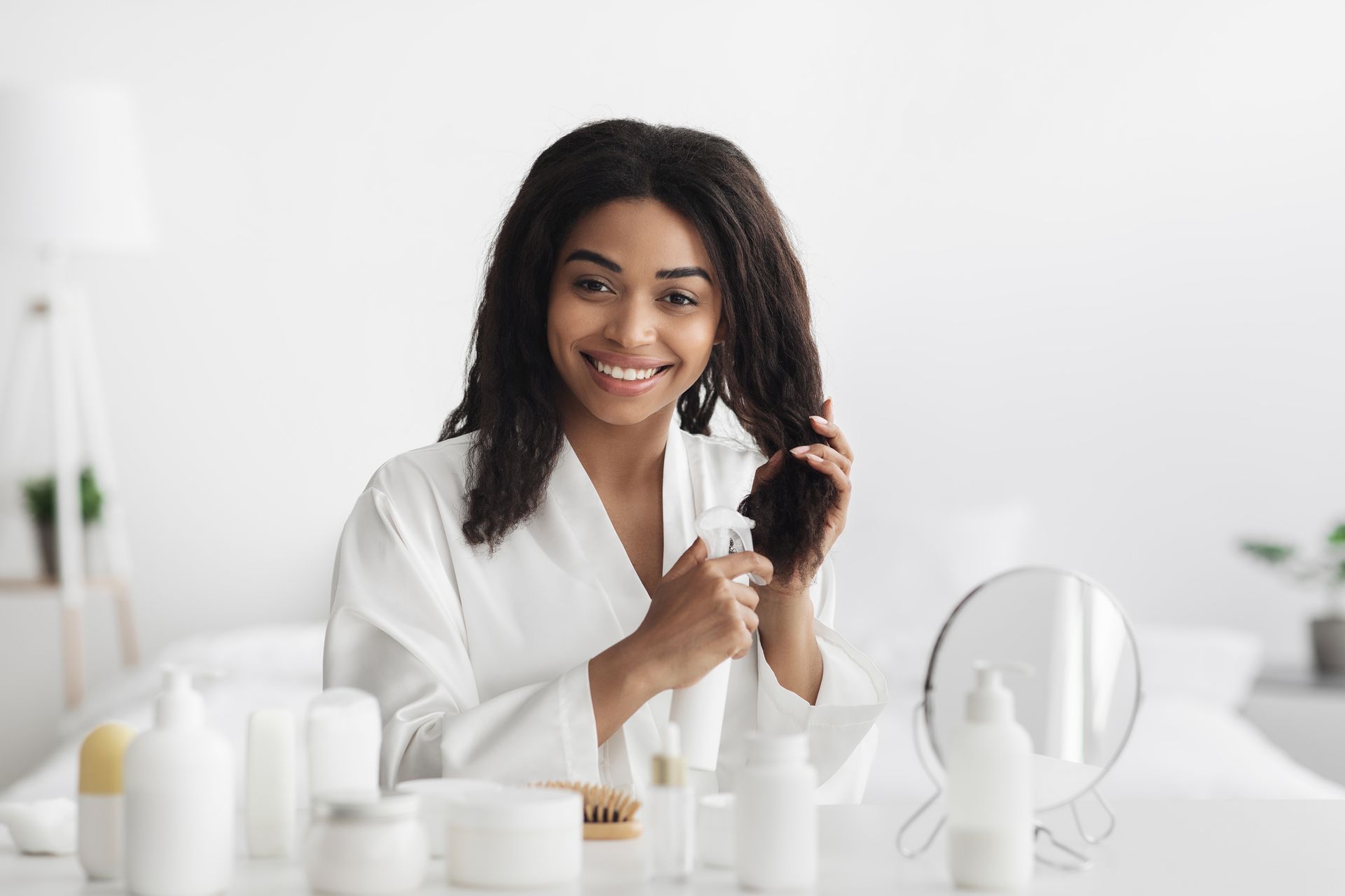 Femme en robe blanche souriante, tenant ses cheveux et un flacon pulvérisateur, entourée de produits de soin pour la peau.