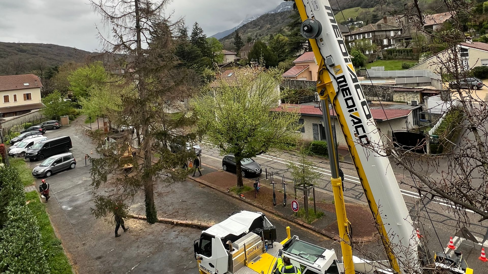 Une nacelle élévatrice soulève des travailleurs au-dessus d'un parking détrempé, encombré d'arbres et de bâtiments après la pluie.