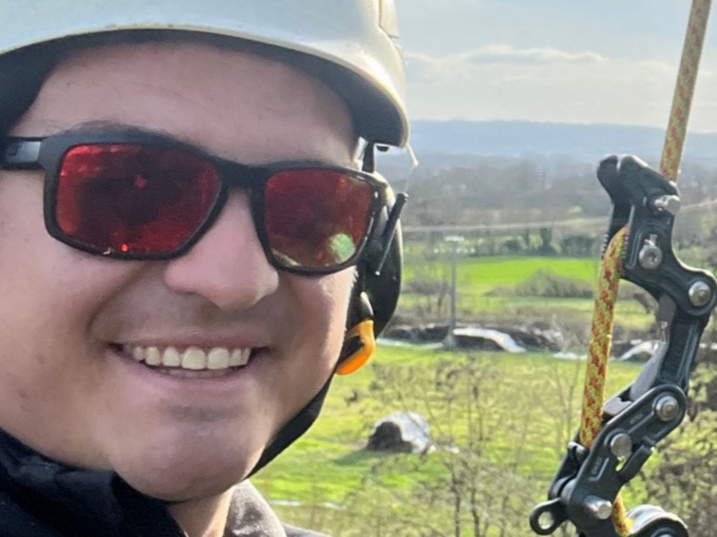 Une personne souriante, portant un casque et des lunettes de soleil rouges, se trouve à l'extérieur, avec un champ vert et des collines en arrière-plan.