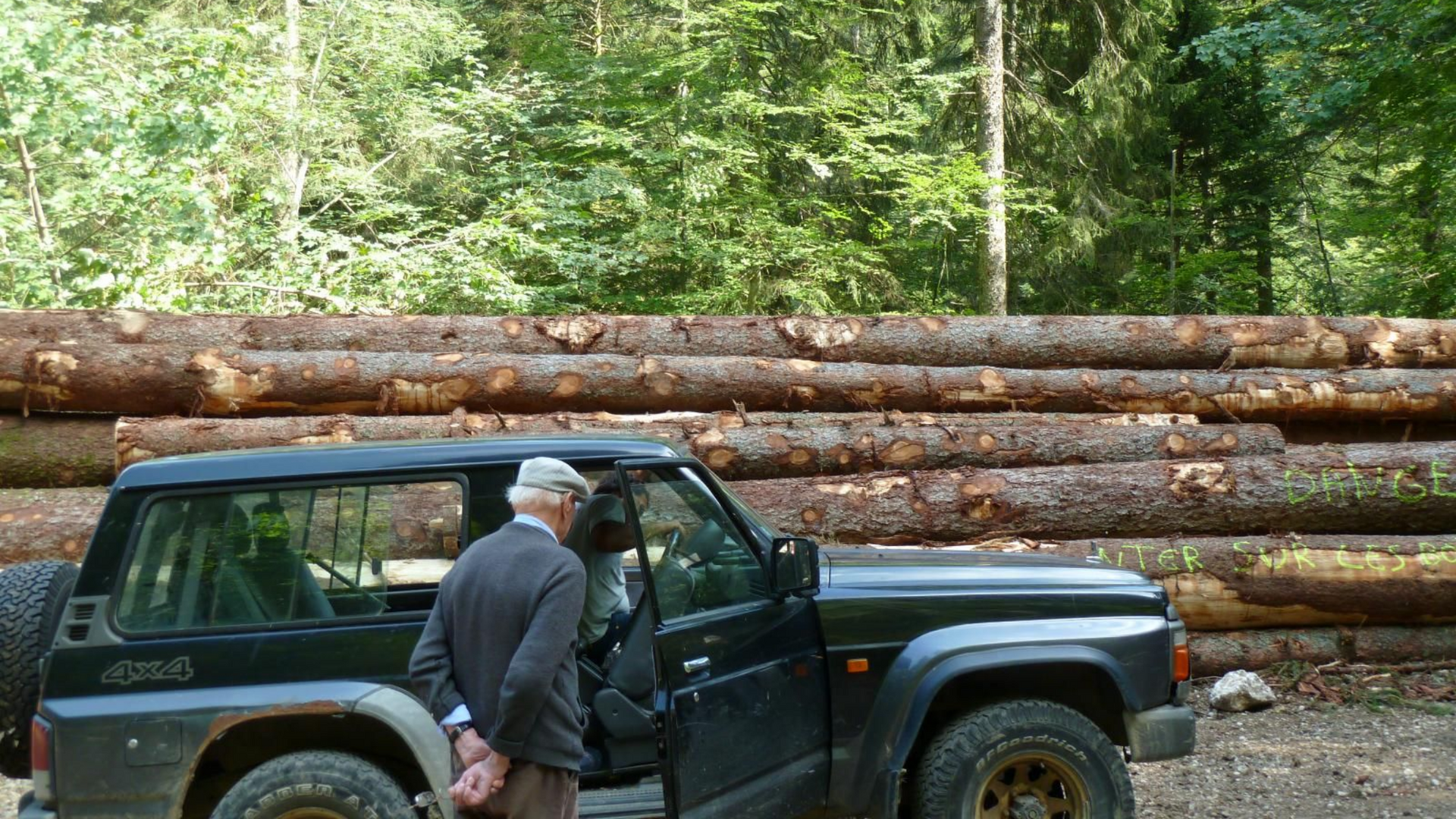 Un homme à côté d'un SUV noir, garé près d'une pile de bûches dans une forêt.