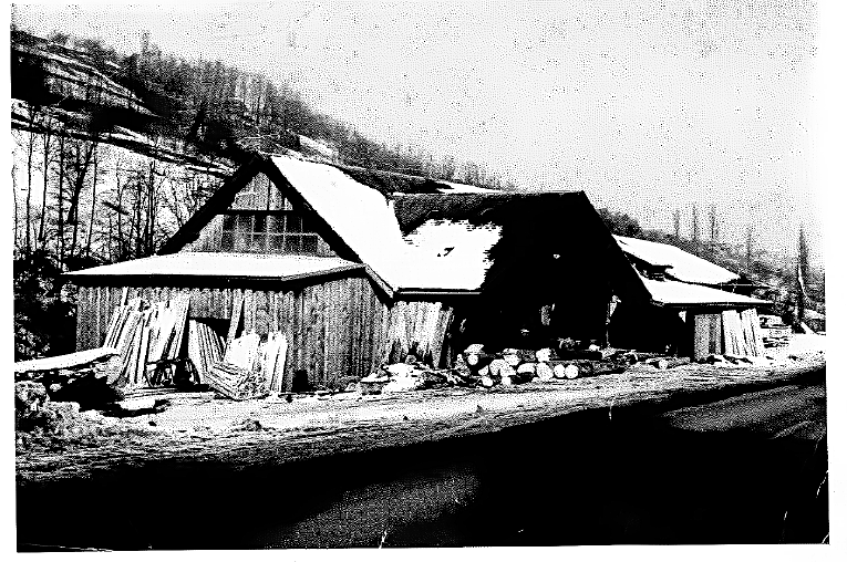 Bâtiment en bois au toit enneigé ; rondins et flanc de colline en arrière-plan.