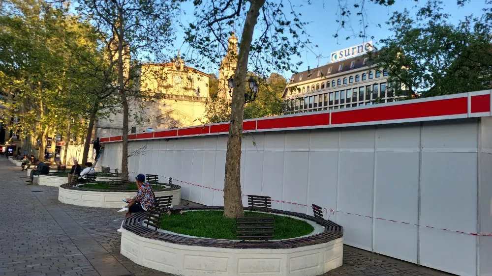 Plaza al aire libre con bancos, árboles y un largo muro blanco con una franja roja frente a los edificios.