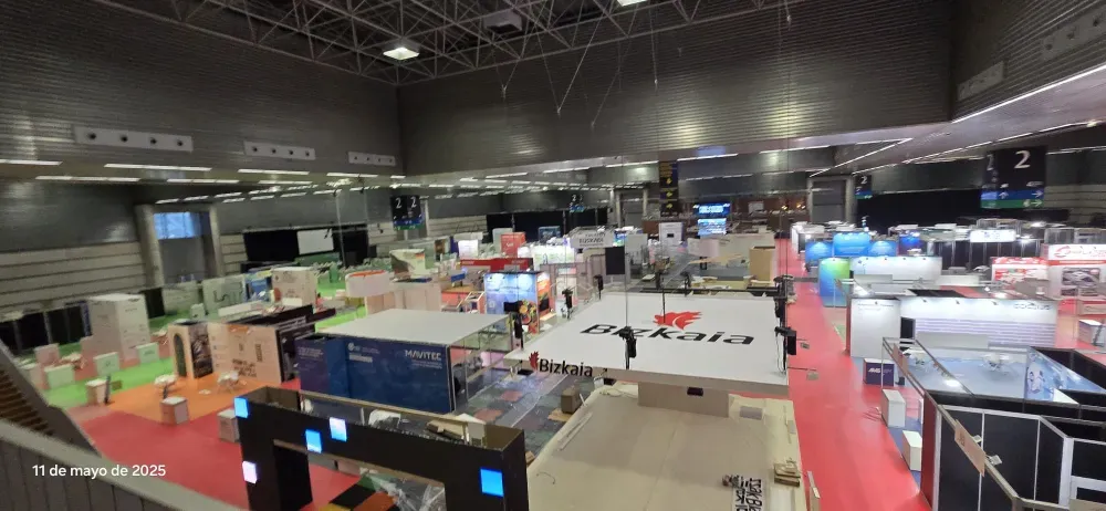 Amplia sala de exposiciones interior con stands, pancartas y algunas personas paseando bajo luces brillantes.