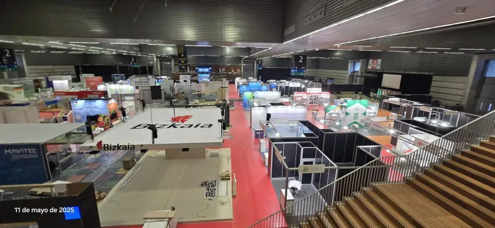 Salón de eventos interior con pasillo alfombrado de rojo, mesas, sillas e iluminación cenital bajo un techo oscuro.
