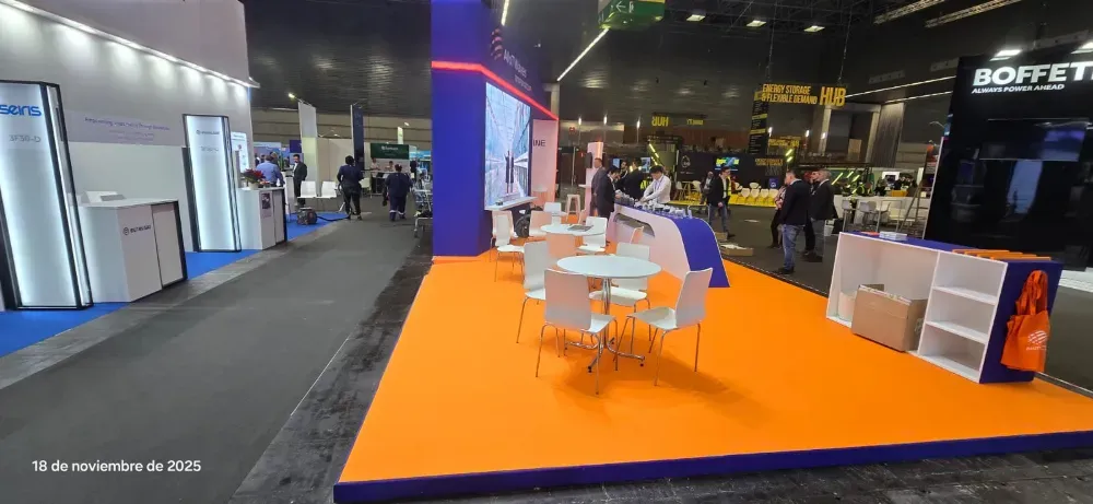 Stand de feria comercial con moqueta naranja, zona de estar y expositores en un gran pabellón de exposiciones.