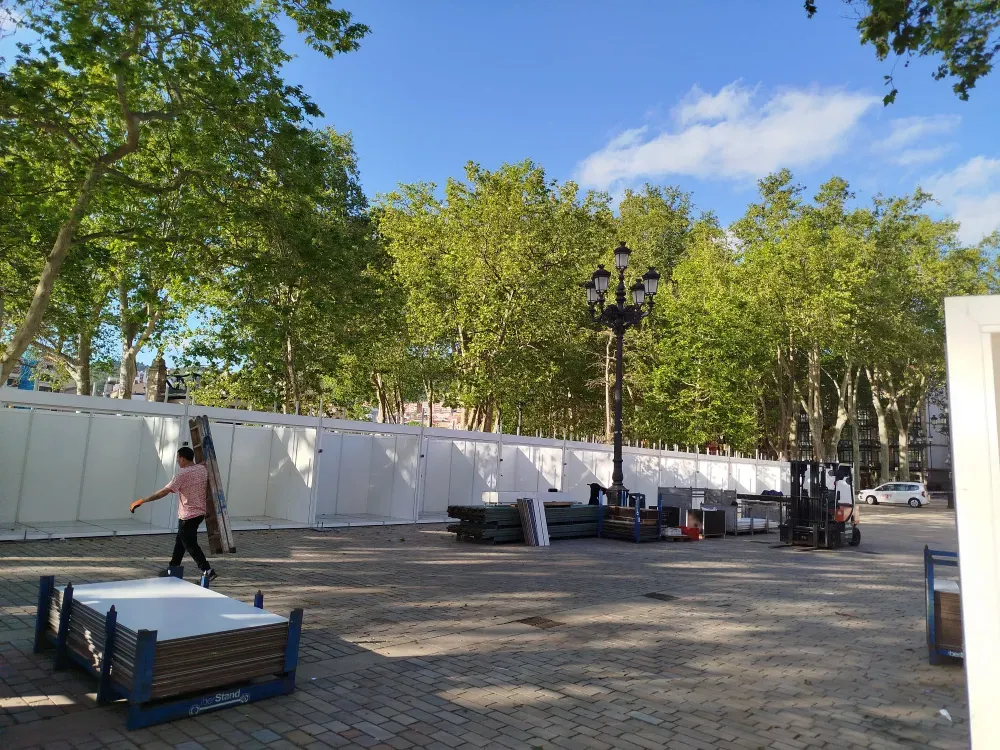 Obra al aire libre con valla blanca, árboles, palés apilados y una persona caminando por una zona pavimentada.