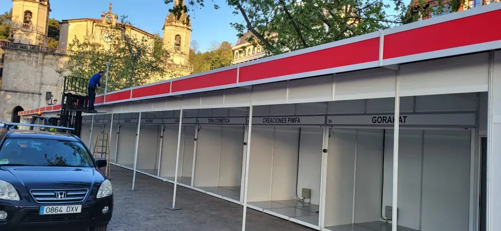 Puestos de mercado vacíos de color rojo y blanco junto a una calle con un coche azul, bajo los árboles y el campanario de una iglesia.