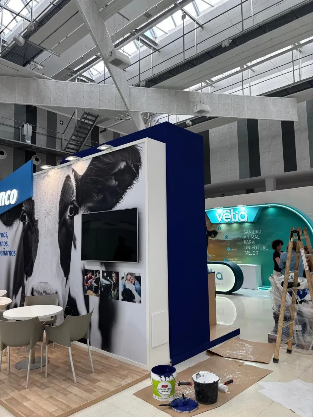 Stand de exposición con paneles blancos y azules, asientos y gráficos de perros grandes en un luminoso salón de convenciones.