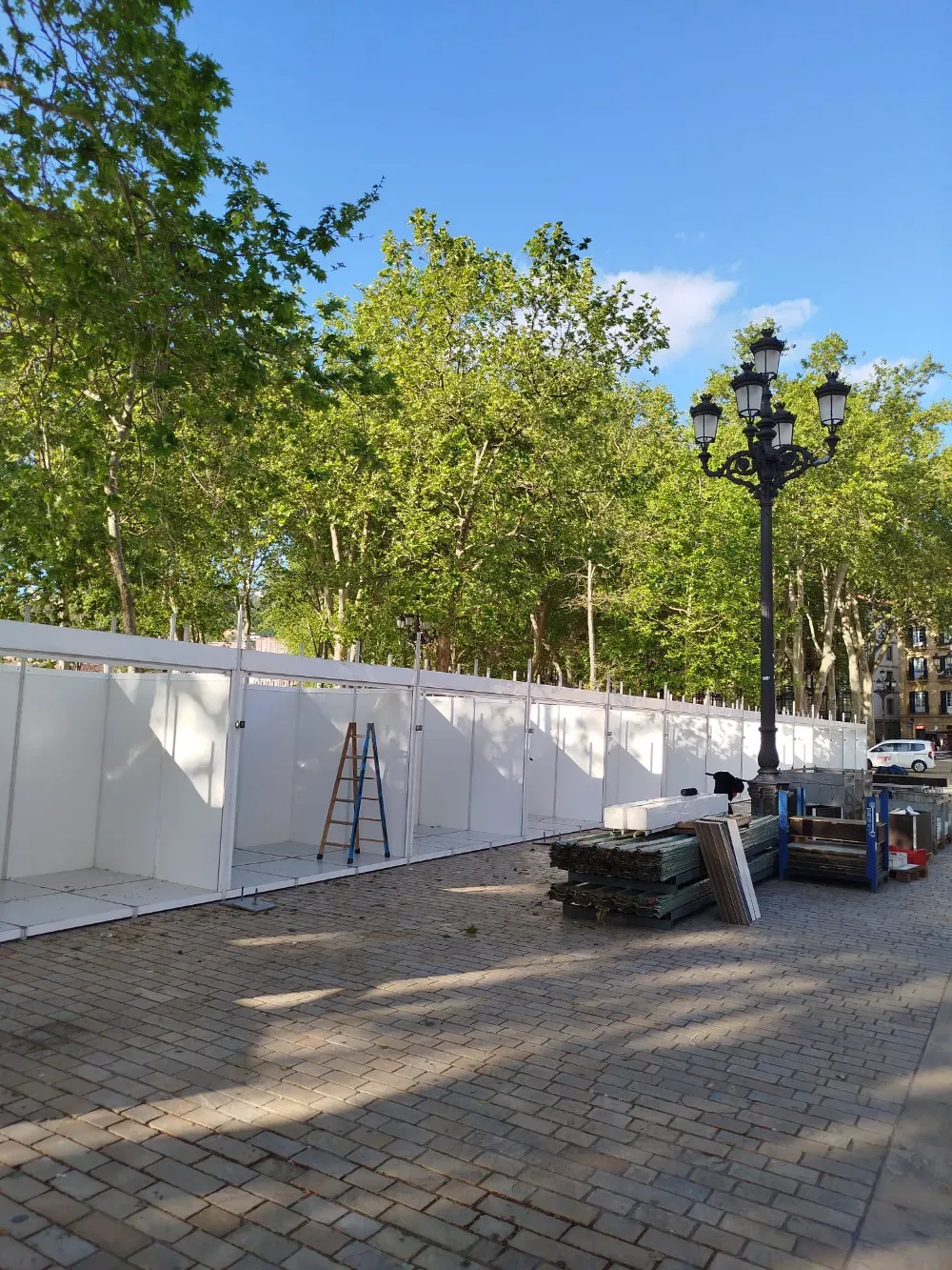Obra en construcción con vallas temporales blancas, escaleras, árboles y un camino empedrado bajo un cielo azul.
