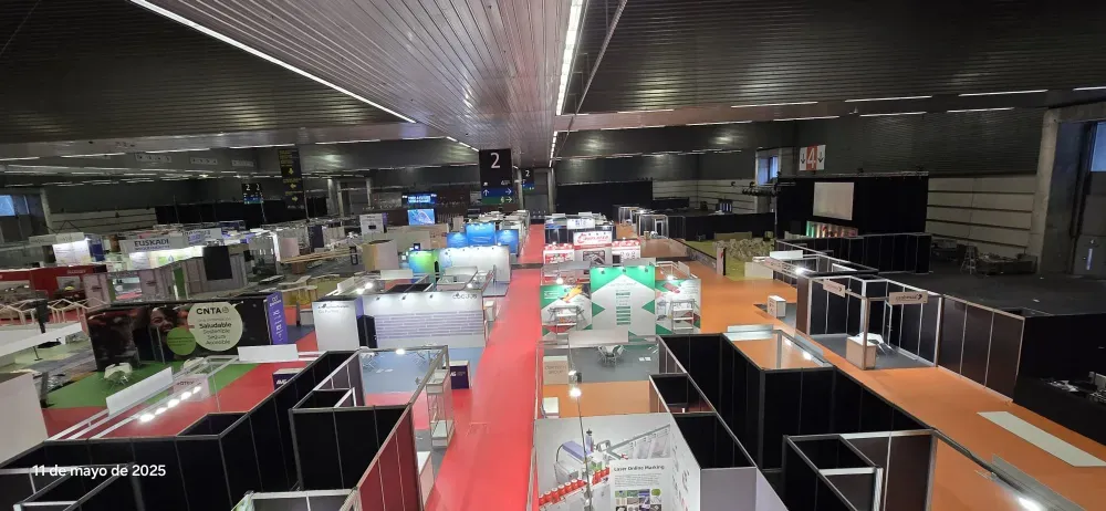Amplia sala de exposiciones interior con filas de stands negros, pasillos alfombrados de rojo e iluminación cenital.