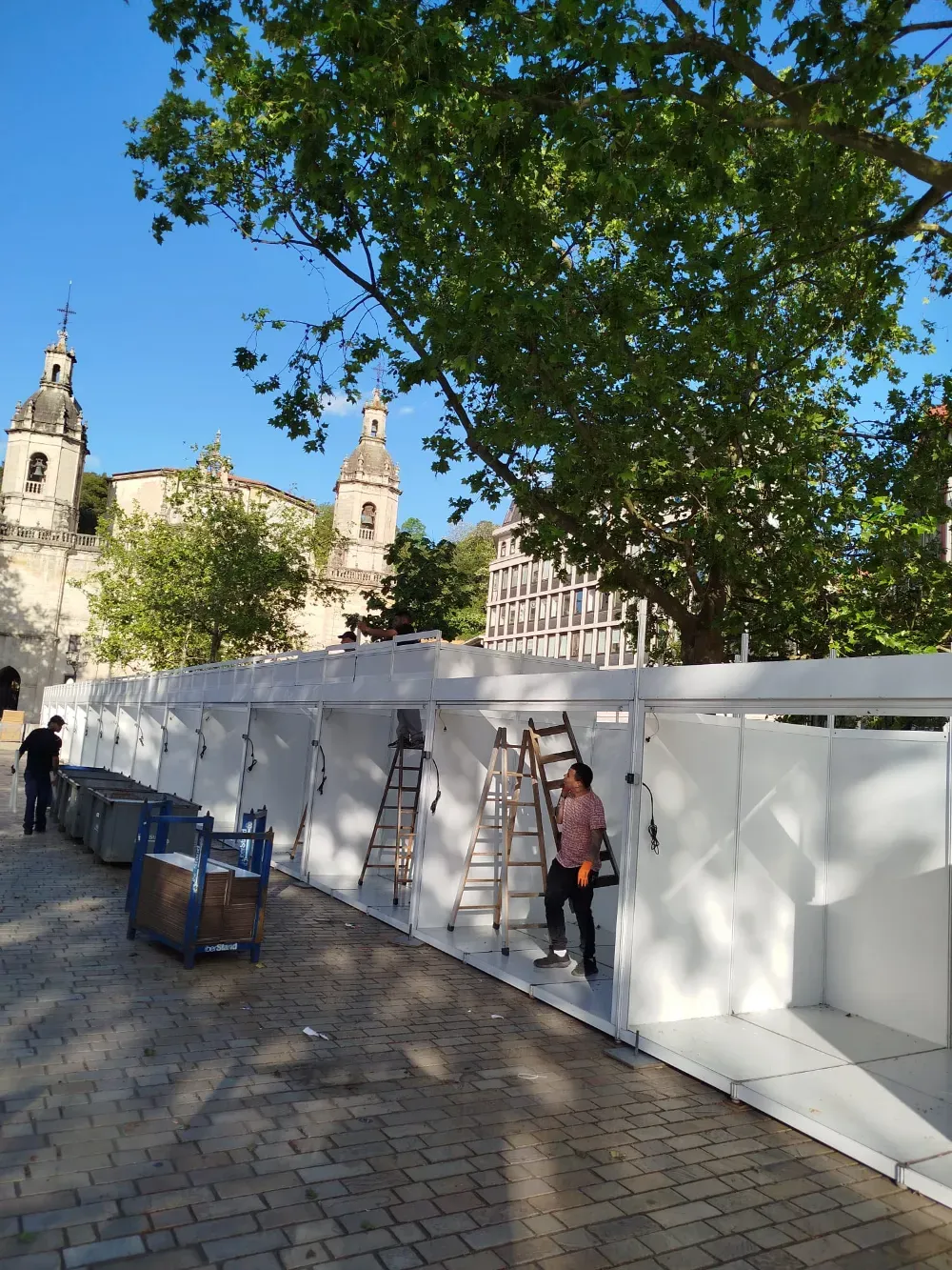 Puestos de mercado blancos bajo un gran árbol en una plaza soleada con un edificio histórico al fondo.