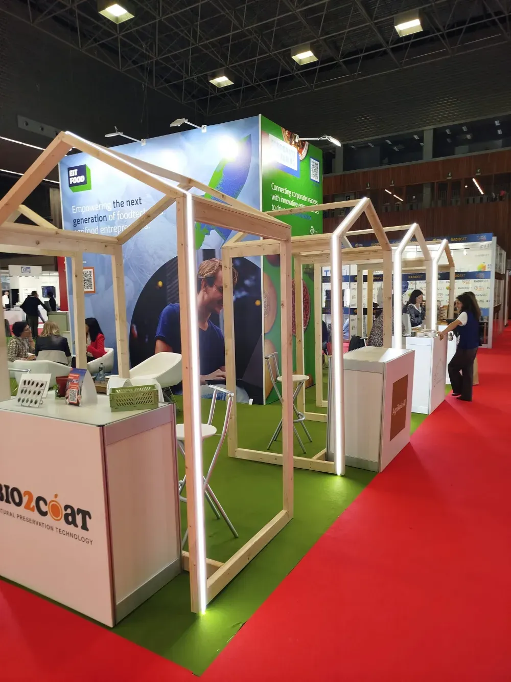 Exhibidores de stand de feria comercial con marcos de madera y paneles verdes y blancos sobre un piso de exposición alfombrado de rojo.