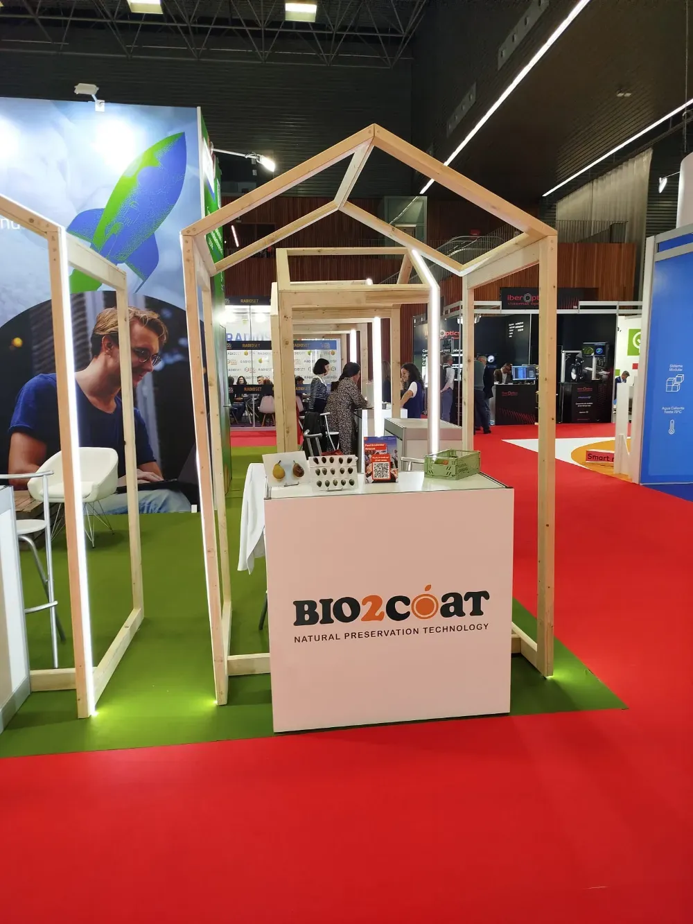 Stand de Bio2coat en la feria, con estructura de madera sobre una alfombra roja, suelo verde y mostrador blanco.