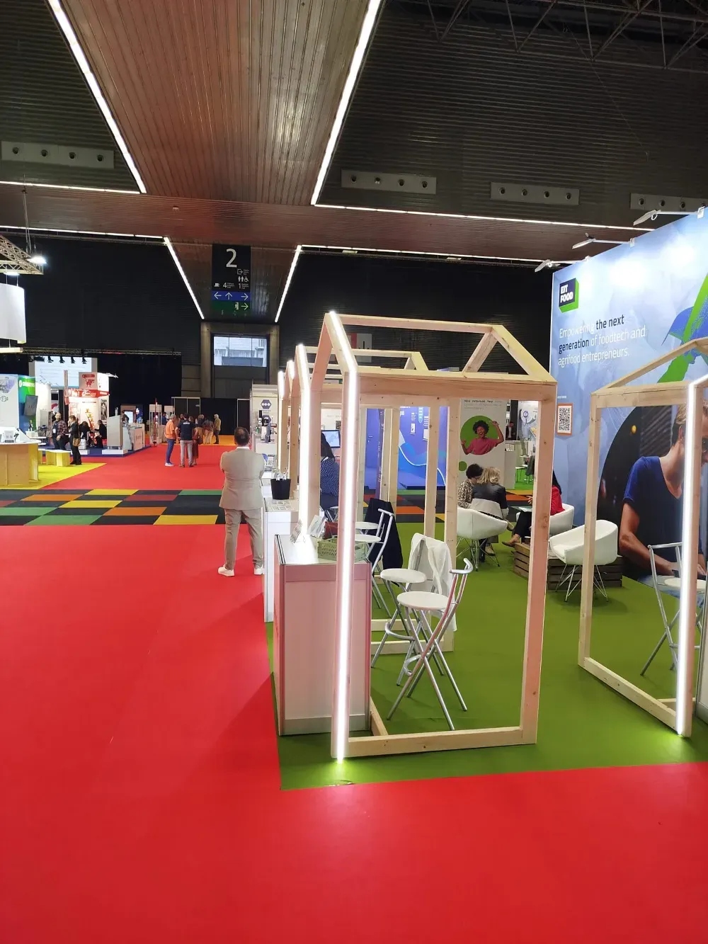 Pabellón de exposiciones interior con alfombra roja y stands de madera iluminados con luces brillantes.