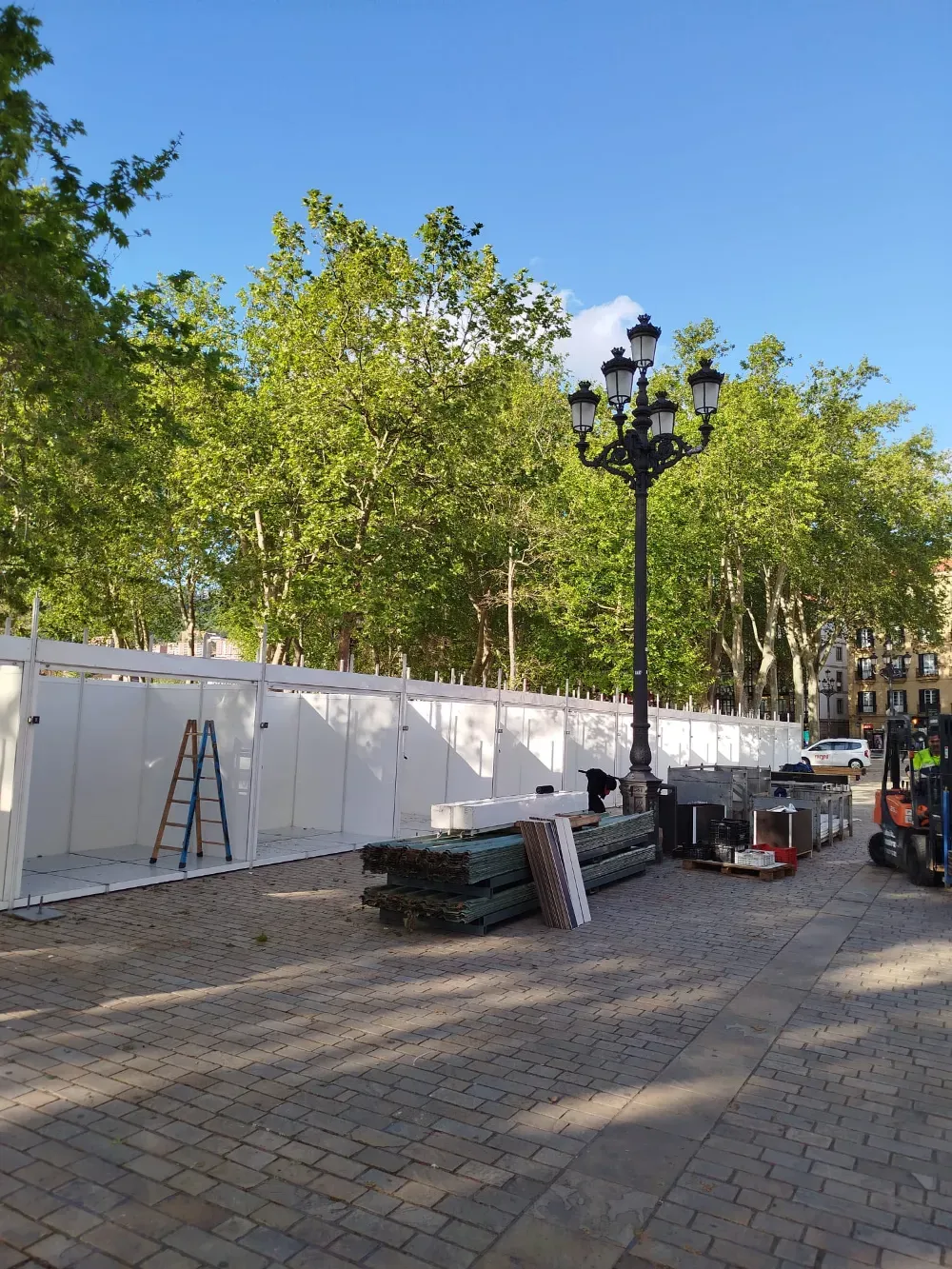 Escena callejera al aire libre con vallas de construcción blancas, árboles, una farola y neumáticos apilados en una calle empedrada.