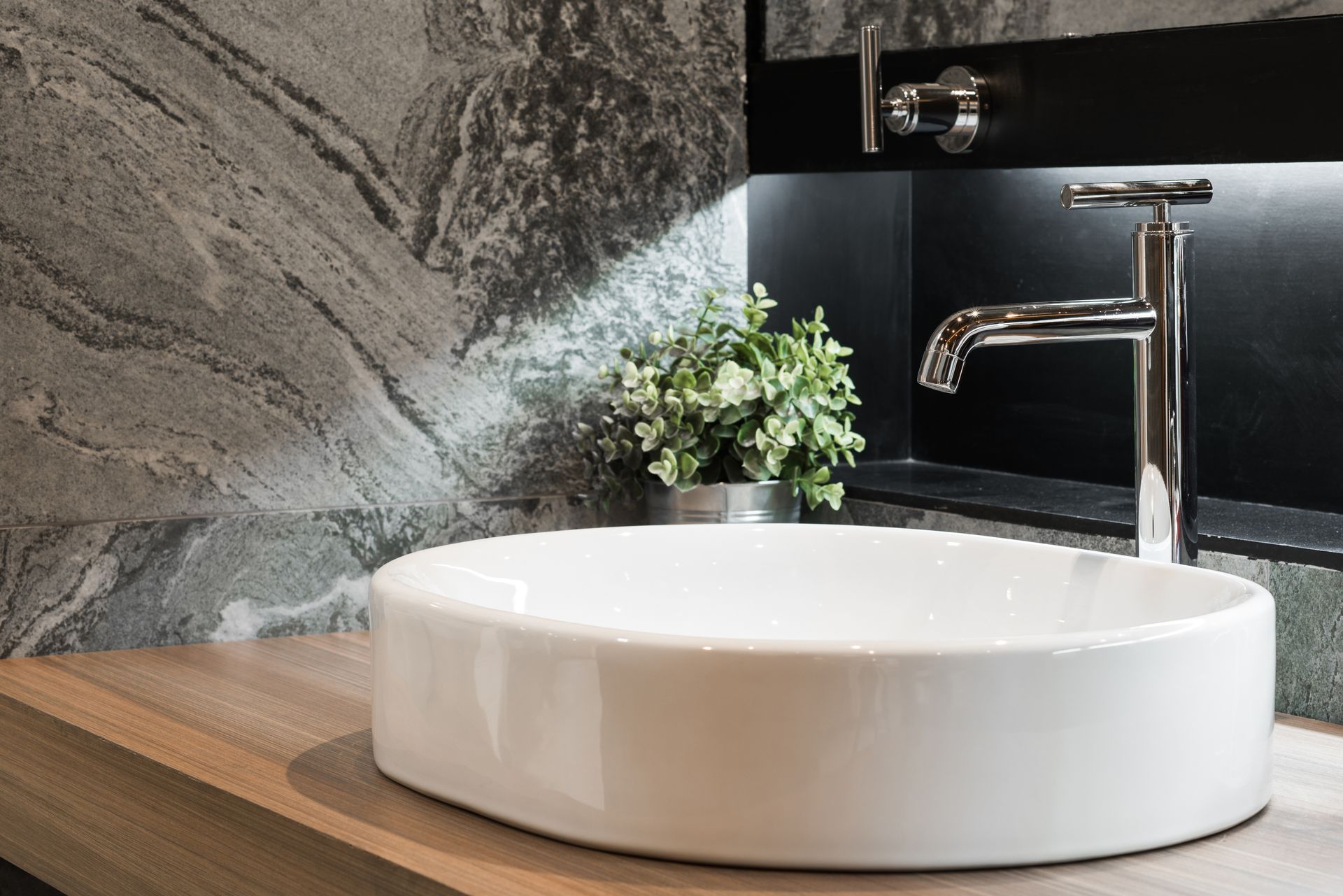 Weißes ovales Waschbecken mit Chromhahn, Grünpflanzen und Marmorwand.