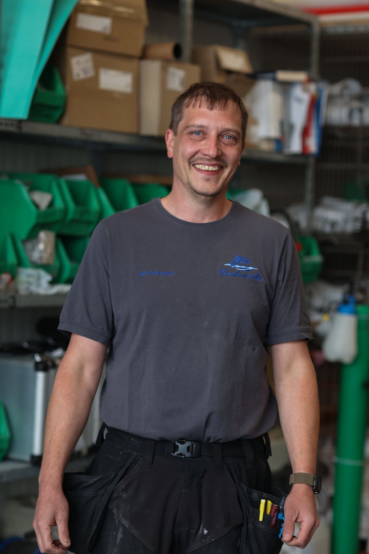 Ein Mann in einem grauen T-Shirt und schwarzen Hosen lächelt in einer Garage.