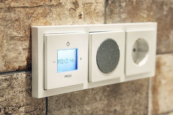 Ein weißer Thermostat ist neben einem Lautsprecher an einer Ziegelwand angebracht.